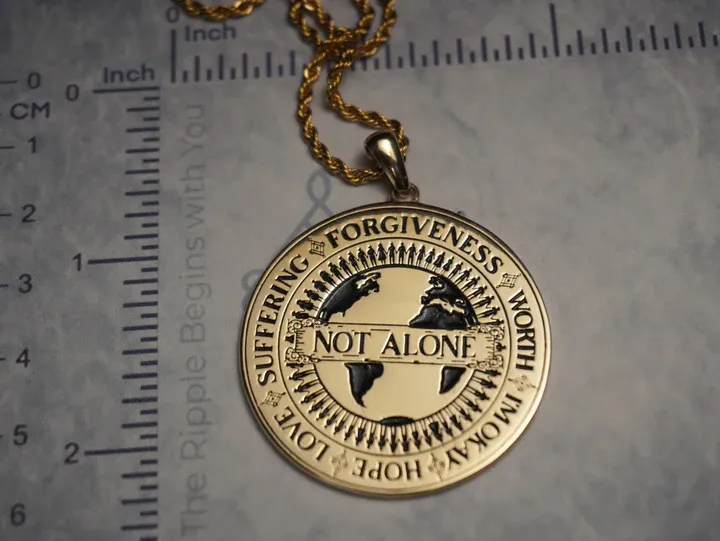 A gold coin necklace placed on a measuring scale showing the "Not Alone" design featuring Ethereal font in use