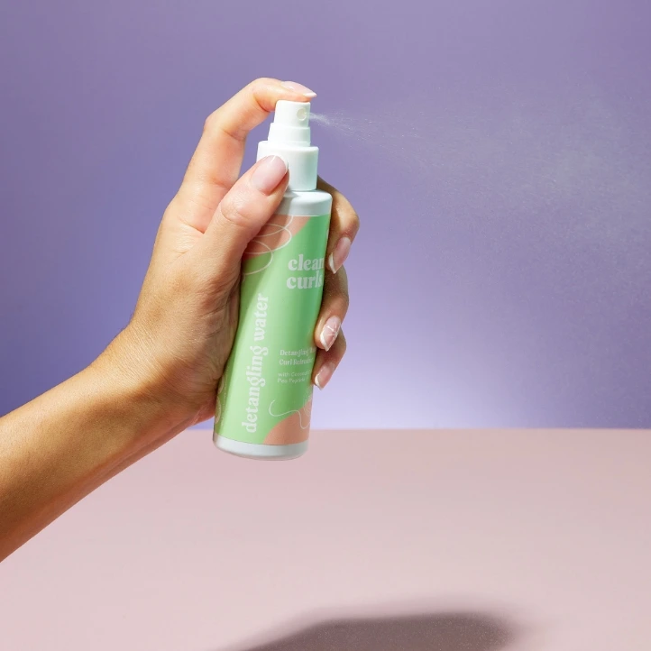 Hand holding a spray bottle of detangling water against a purple background featuring Asikue font in use